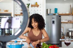 profesional hermosa contento joven mujer es blogging para su cocina canal acerca de sano vivo en el cocina de su hogar y mirando en cámara en un trípode foto