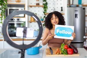 Happy african american woman vlogger broadcasting live video online while cooking food in kitchen at home holding follow sign photo