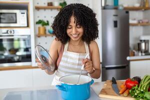 Happy African American Woman Cooking Tasting Dinner In A Pot Standing In Modern Kitchen At Home. Housewife Preparing Healthy Food Smiling To Camera. Household And Nutrition. Dieting Recipes Concept photo