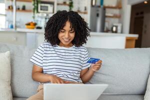 Young Woman On Sofa Shopping Online With Debit Card. Beautiful African woman using laptop computer for online shopping at home photo