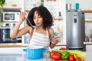 Happy Young Woman Cooking Tasting Dinner In A Pot Standing In Modern Kitchen At Home. Housewife Preparing Healthy Food Smiling . Household And Nutrition. Dieting Recipes Concept photo