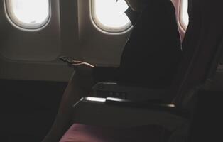 Blonde female tourist checking incoming notification on smartphone sitting on seat of airplane with netbook.Young businesswoman share media from telephone on laptop computer during plane flight photo