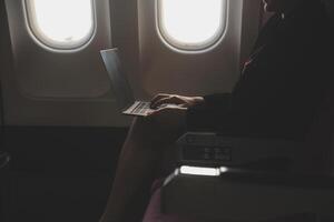 Blonde female tourist checking incoming notification on smartphone sitting on seat of airplane with netbook.Young businesswoman share media from telephone on laptop computer during plane flight photo