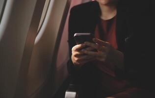 Blonde female tourist checking incoming notification on smartphone sitting on seat of airplane with netbook.Young businesswoman share media from telephone on laptop computer during plane flight photo