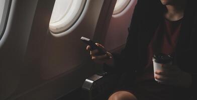 Blonde female tourist checking incoming notification on smartphone sitting on seat of airplane with netbook.Young businesswoman share media from telephone on laptop computer during plane flight photo