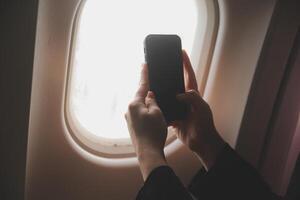 Blonde female tourist checking incoming notification on smartphone sitting on seat of airplane with netbook.Young businesswoman share media from telephone on laptop computer during plane flight photo