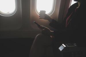 Blonde female tourist checking incoming notification on smartphone sitting on seat of airplane with netbook.Young businesswoman share media from telephone on laptop computer during plane flight photo
