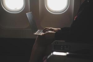 Blonde female tourist checking incoming notification on smartphone sitting on seat of airplane with netbook.Young businesswoman share media from telephone on laptop computer during plane flight photo