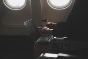 Blonde female tourist checking incoming notification on smartphone sitting on seat of airplane with netbook.Young businesswoman share media from telephone on laptop computer during plane flight photo