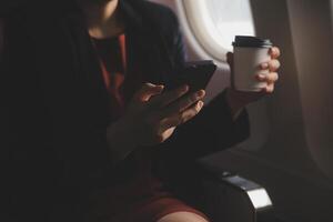 Blonde female tourist checking incoming notification on smartphone sitting on seat of airplane with netbook.Young businesswoman share media from telephone on laptop computer during plane flight photo