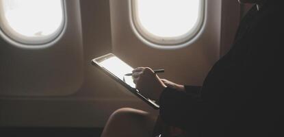 Blonde female tourist checking incoming notification on smartphone sitting on seat of airplane with netbook.Young businesswoman share media from telephone on laptop computer during plane flight photo