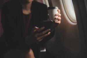 Blonde female tourist checking incoming notification on smartphone sitting on seat of airplane with netbook.Young businesswoman share media from telephone on laptop computer during plane flight photo