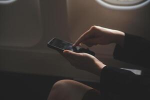 Blonde female tourist checking incoming notification on smartphone sitting on seat of airplane with netbook.Young businesswoman share media from telephone on laptop computer during plane flight photo