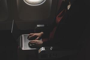 Blonde female tourist checking incoming notification on smartphone sitting on seat of airplane with netbook.Young businesswoman share media from telephone on laptop computer during plane flight photo