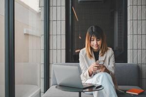 Happy young asian woman using mobile phone while sitting on safa at home with laptop computer. photo