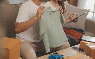 Two Asian blogger showing clothes in front of the camera to recording vlog video live streaming at her shop. Online Shopping Cart notion. on sofa at home photo