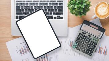 A top-down view of a professional's workspace with a tablet, a laptop, financial documents, a calculator, and a cup of coffee next to a lush potted plant, all laid out on a wooden surface. photo