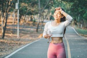 A radiant fitness enthusiast wipes sweat from her brow while holding a water bottle during a jogging break, set against a serene park backdrop, embodying active wellness. photo