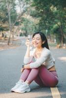 Sport fitness concept, A cheerful young woman sitting on a forest road takes a water break after a run, enjoying her fitness routine outdoors. photo