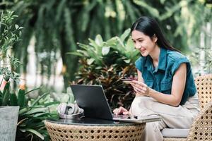 A smiling woman sits comfortably outdoors with a laptop and headphones, holding a credit card, ready to complete an online transaction in a tranquil garden environment. photo