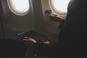 Blonde female tourist checking incoming notification on smartphone sitting on seat of airplane with netbook.Young businesswoman share media from telephone on laptop computer during plane flight photo