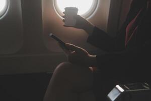 Blonde female tourist checking incoming notification on smartphone sitting on seat of airplane with netbook.Young businesswoman share media from telephone on laptop computer during plane flight photo