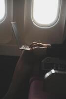 Blonde female tourist checking incoming notification on smartphone sitting on seat of airplane with netbook.Young businesswoman share media from telephone on laptop computer during plane flight photo