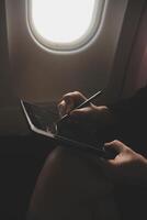 Blonde female tourist checking incoming notification on smartphone sitting on seat of airplane with netbook.Young businesswoman share media from telephone on laptop computer during plane flight photo