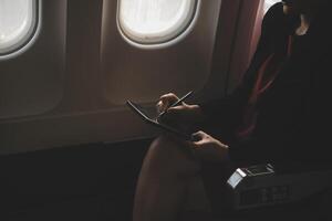 Blonde female tourist checking incoming notification on smartphone sitting on seat of airplane with netbook.Young businesswoman share media from telephone on laptop computer during plane flight photo
