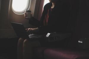 Blonde female tourist checking incoming notification on smartphone sitting on seat of airplane with netbook.Young businesswoman share media from telephone on laptop computer during plane flight photo