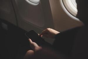 Blonde female tourist checking incoming notification on smartphone sitting on seat of airplane with netbook.Young businesswoman share media from telephone on laptop computer during plane flight photo