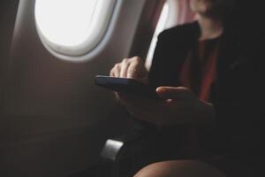 Blonde female tourist checking incoming notification on smartphone sitting on seat of airplane with netbook.Young businesswoman share media from telephone on laptop computer during plane flight photo
