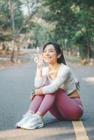 A cheerful young woman in sportswear takes a refreshing break, holding a water bottle while sitting on a park path, exemplifying healthy lifestyle and hydration. photo