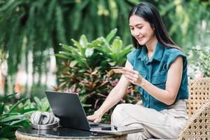 A smiling woman sits comfortably outdoors with a laptop and headphones, holding a credit card, ready to complete an online transaction in a tranquil garden environment. photo