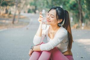 A young woman enjoys a peaceful moment, holding a clear water bottle with a gentle smile, as she takes a hydration break during an outdoor workout, wearing a smartwatch, wireless earbuds. photo