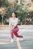 A smiling woman takes a moment to stretch her legs on a running path, wearing pink leggings and white sneakers, with a backdrop of tranquil trees. photo