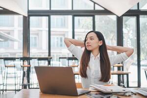 A contemplative woman takes a break, hands behind her head, looking out with a thoughtful gaze, surrounded by her work environment bathed in natural light. photo