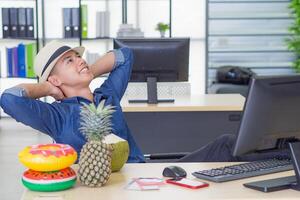 A young man wearing a hat, looking up, placing both hands on the back of the neck and foot on the table for relaxing while sitting on a chair in the office. Holiday coming soon. Holiday concept photo