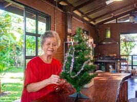 Elderly Asian woman decorating the Christmas tree with white and red ribbons at home. Concept of aged people and Christmas and happy new year festival photo