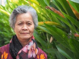 Senior Asian woman with short white hair smiling and looking at the camera while standing in a garden. Space for text. Concept of aged people and healthcare photo