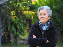 mayor asiático mujer con corto blanco pelo sonriente y mirando a el cámara mientras en pie en un jardín. espacio para texto. concepto de Envejecido personas y cuidado de la salud foto