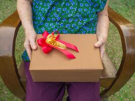 regalo marrón caja con un rojo cinta participación por un mayor mujer mientras sentado en silla. de cerca foto. concepto de Envejecido personas y festival foto