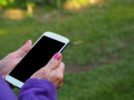 Close-up of wrinkled hands of a senior woman holding a smartphone while standing in a garden. Space for text photo