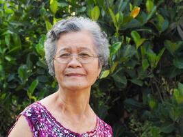 Senior Asian woman with short white hair smiling and looking at the camera while standing in a garden. Space for text. Concept of aged people and healthcare photo