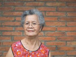 An elderly Asian woman smiling and looking at the camera while standing with a brick wall background. Space for text. Concept of aged people and healthcare photo