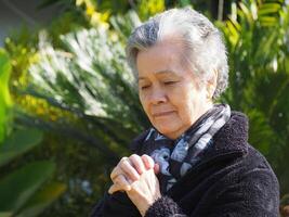 An elderly Asian woman praying and closed eyes while standing in a garden. Space for text. Concept of aged people and healthcare photo