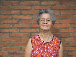 An elderly Asian woman smiling and looking at the camera while standing with a brick wall background. Space for text. Concept of aged people and healthcare photo