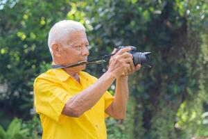 Senior man taking a photo by a digital camera while standing in the park. An elderly Asian man wears a yellow shirt be happy when using a camera. Concept of aged people and photography