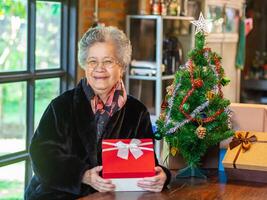 mayor asiático mujer participación un regalo caja y sentado cerca un Navidad árbol a hogar. concepto de Envejecido personas y festival foto