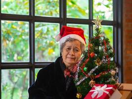 mayor mujer sonriente y mirando a el cámara mientras sentado cerca un Navidad árbol a hogar. concepto de Envejecido personas y Navidad y contento nuevo año festival foto